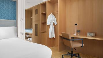 A hotel room with a bed, a wooden wardrobe, a desk with a chair, and a robe hanging on the wardrobe.