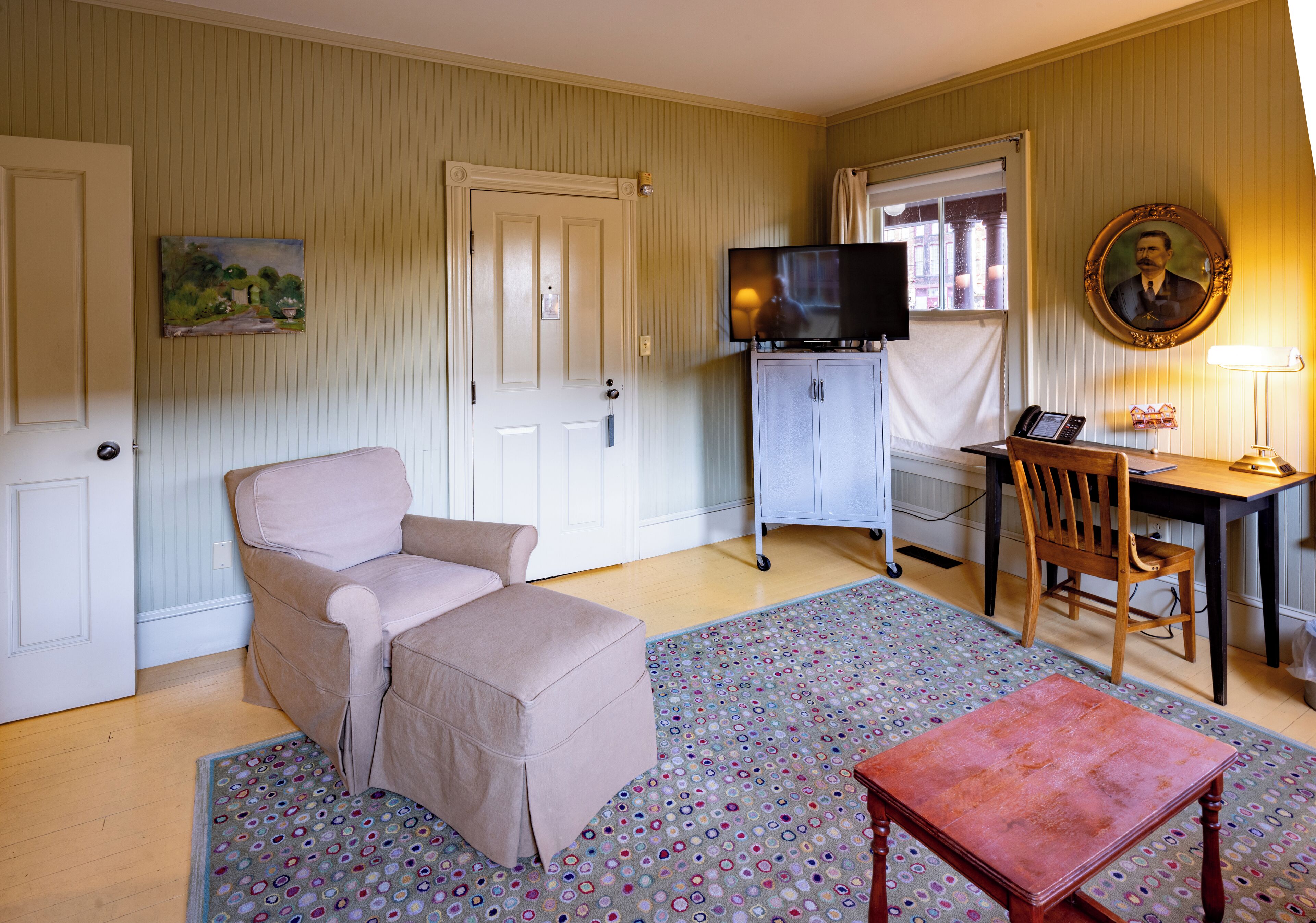 A room with a beige sofa, a small table, a TV on a stand, a desk with a chair, and a patterned rug.