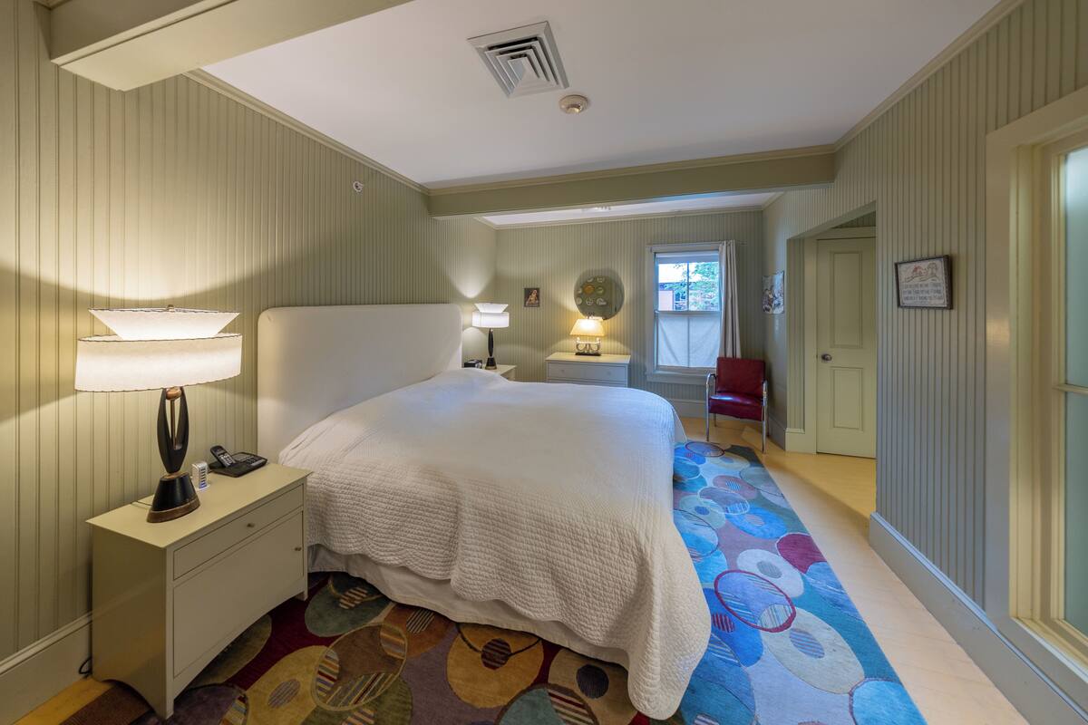 Une chambre d’hôtel avec un grand lit, une table de chevet avec une lampe, une petite table avec une lampe, un tapis à motifs et une porte donnant sur une autre pièce.