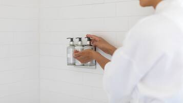 En person i en hvid badekåbe rækker efter en sæbedispenser på en flisebeklædt væg.