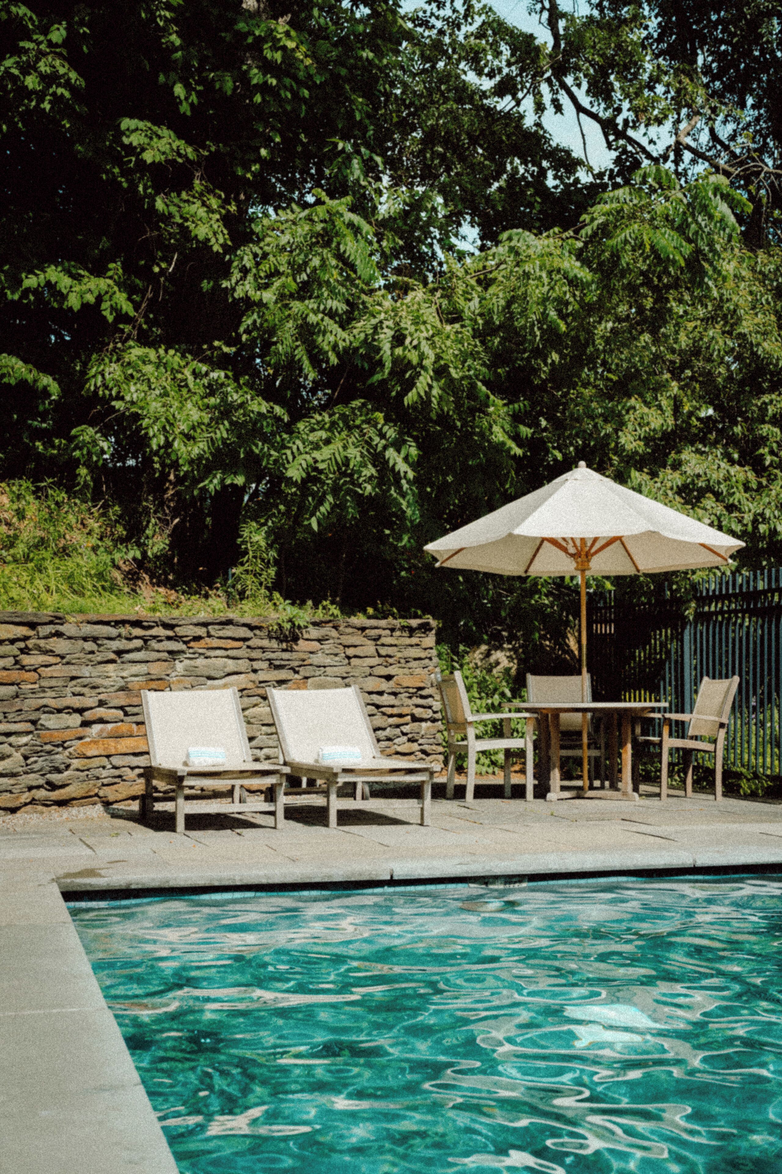 Ein Pool mit Liegestühlen und Sonnenschirm, umgeben von einer Steinmauer und Bäumen.