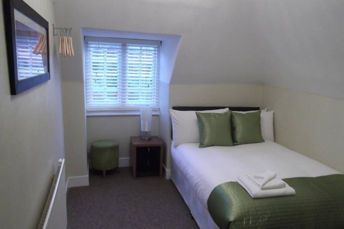 Une chambre avec un lit simple, une table de chevet, une lampe, une fenêtre avec des stores et un tableau encadré au mur.