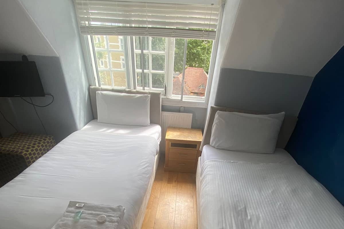 Une petite chambre d’hôtel, composée d’une seule pièce, avec deux lits, un plancher en bois, une fenêtre avec des stores et un radiateur.