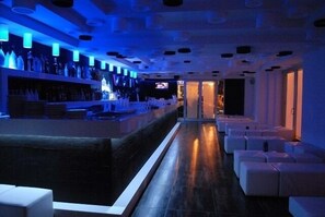 Eine Bar mit einer langen Theke, Regalen voller Flaschen und einem Fernsehbildschirm. Weiße Sitzgelegenheiten säumen die Wände.