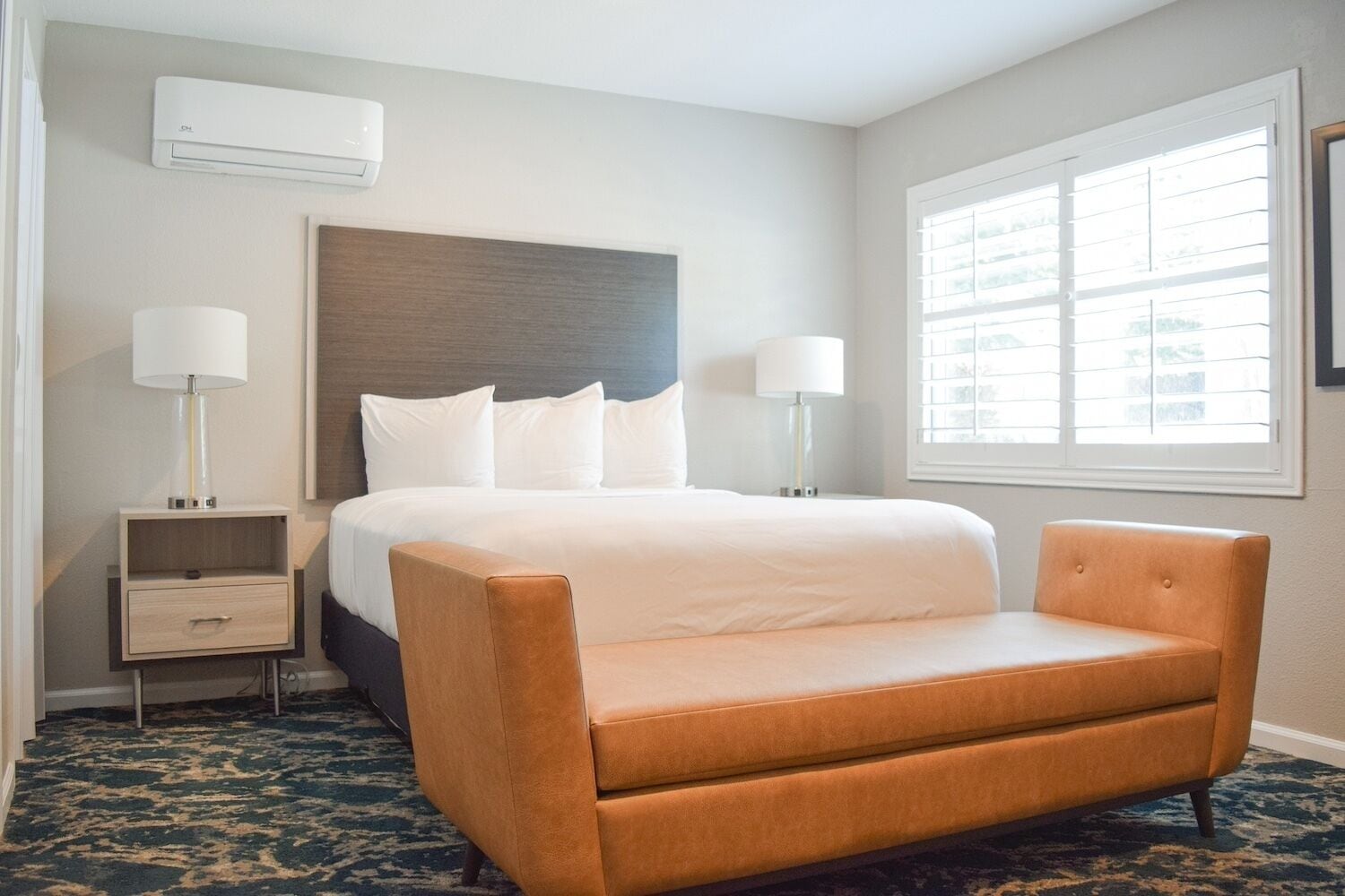 A hotel room with a bed, two bedside lamps, a nightstand, a dresser, and a window with blinds.