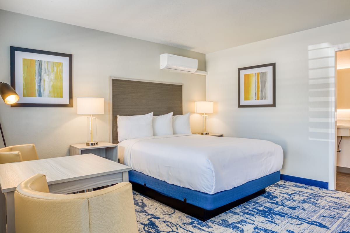 A hotel room with a large bed, a desk, and two chairs. There are two bedside tables with lamps, a wall-mounted air conditioner, and two framed pictures on the walls.