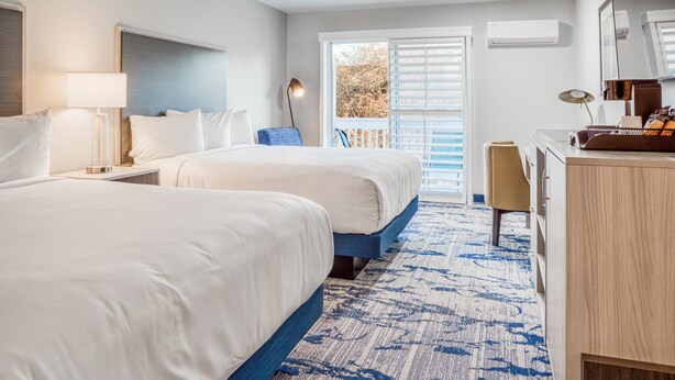 Ein Hotelzimmer mit zwei Betten, einem Schreibtisch, einem Stuhl, einer Lampe und einem großen Fenster mit Jalousien.