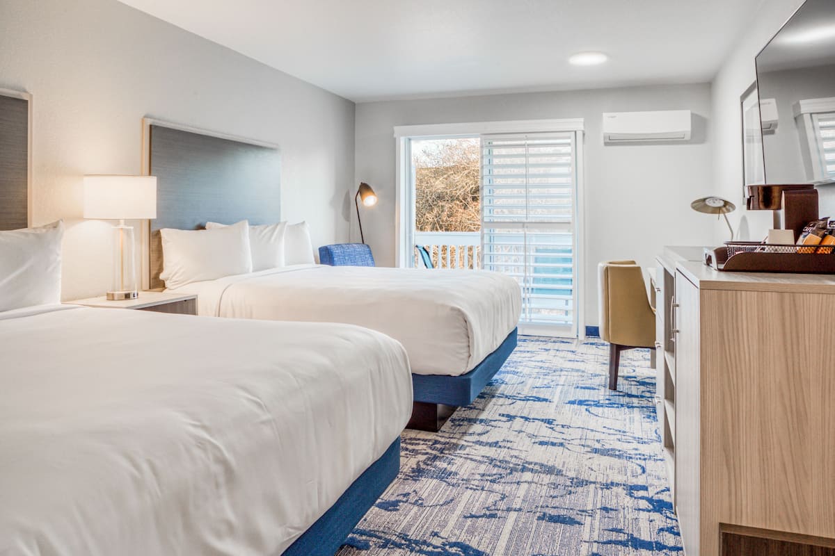 A hotel room with two beds, a desk, a chair, a lamp, and a large window with blinds.