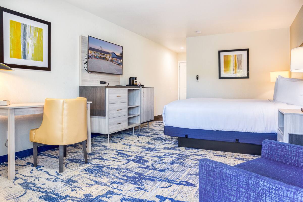 A hotel room with a bed, a desk, a chair, a dresser, and artwork on the walls.