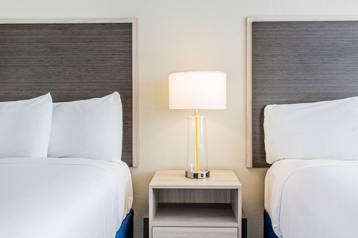 A hotel room with two beds, each with white bedding and a wooden headboard. A bedside table with a lamp is between the beds.