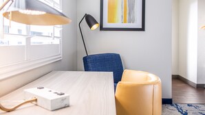 Ein modernes Zimmer mit einem Holztisch, einem blauen Stuhl, einem gelben Hocker, einer schwarzen Stehlampe und einem gerahmten abstrakten Gemälde an der Wand.