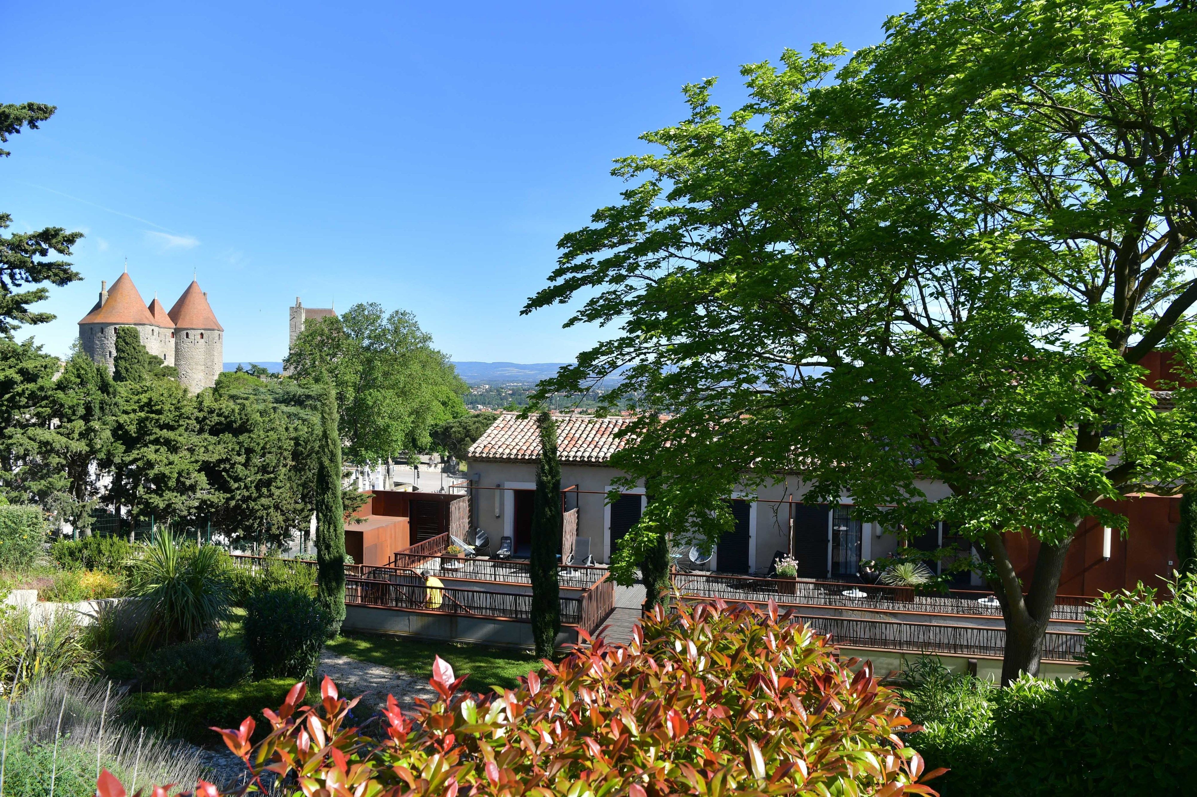 A castle with two towers, surrounded by lush greenery and a modern building with a tiled roof.