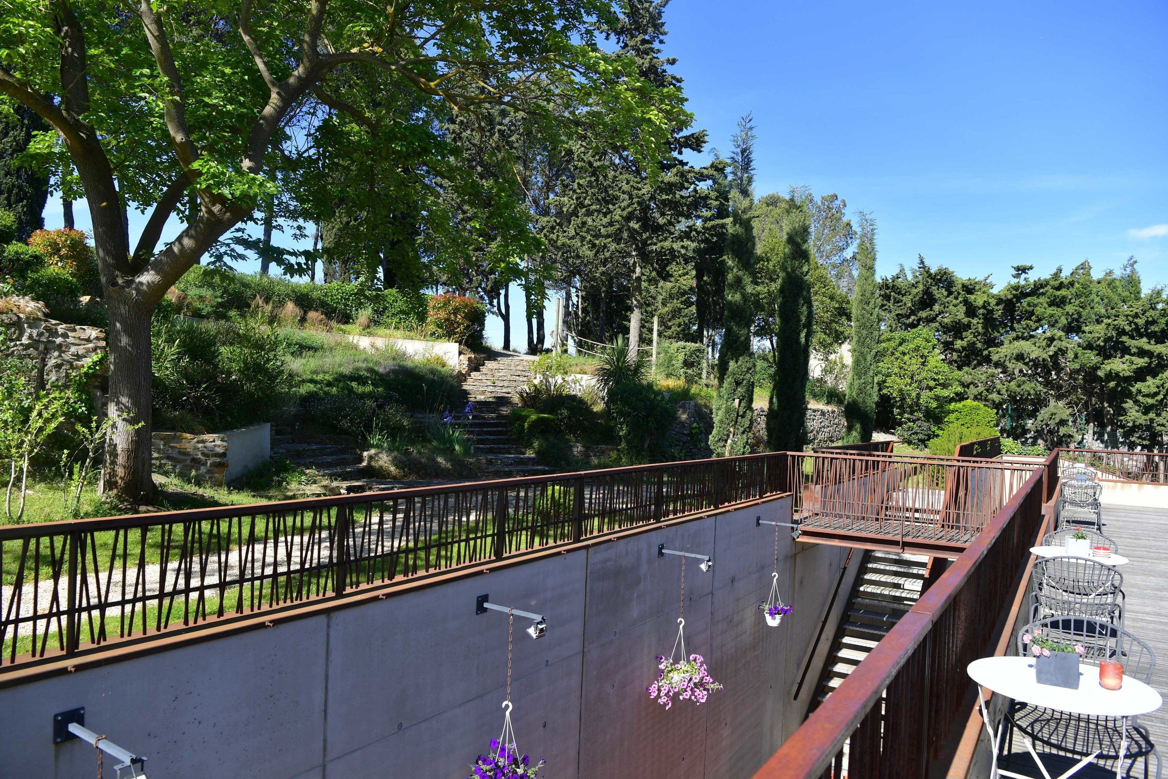 A balcony with hanging flower baskets, a metal railing, and a view of a landscaped garden with a stone wall and stairs leading down.