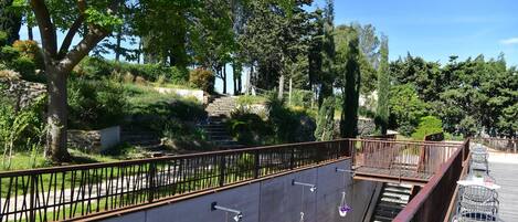 A balcony with hanging flower baskets, a metal railing, and a view of a landscaped garden with a stone wall and stairs leading down.
