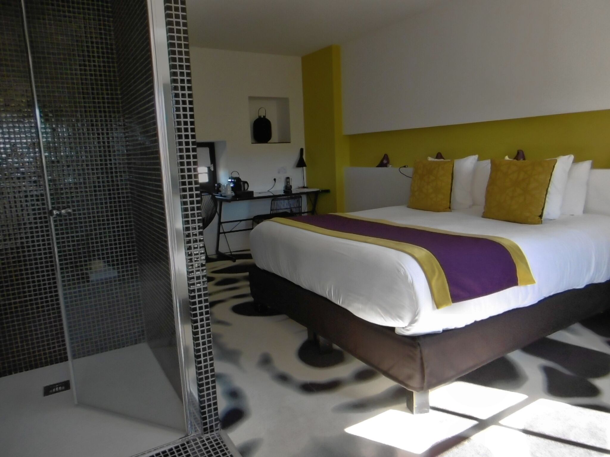 Ett hotellrum med en säng, ett skrivbord, en stol och en duschhörna.