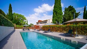 Une piscine à l’eau d’un bleu limpide, entourée de chaises longues et de parasols, et une clôture bordée de grands arbres en arrière-plan.