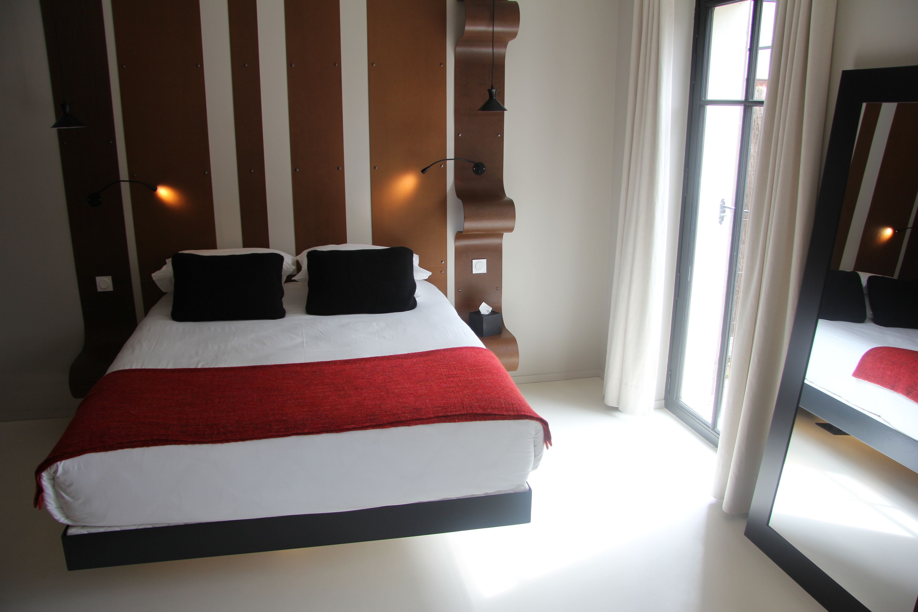 A hotel room with a large bed, a wooden headboard, a window with curtains, and a mirror on the wall.