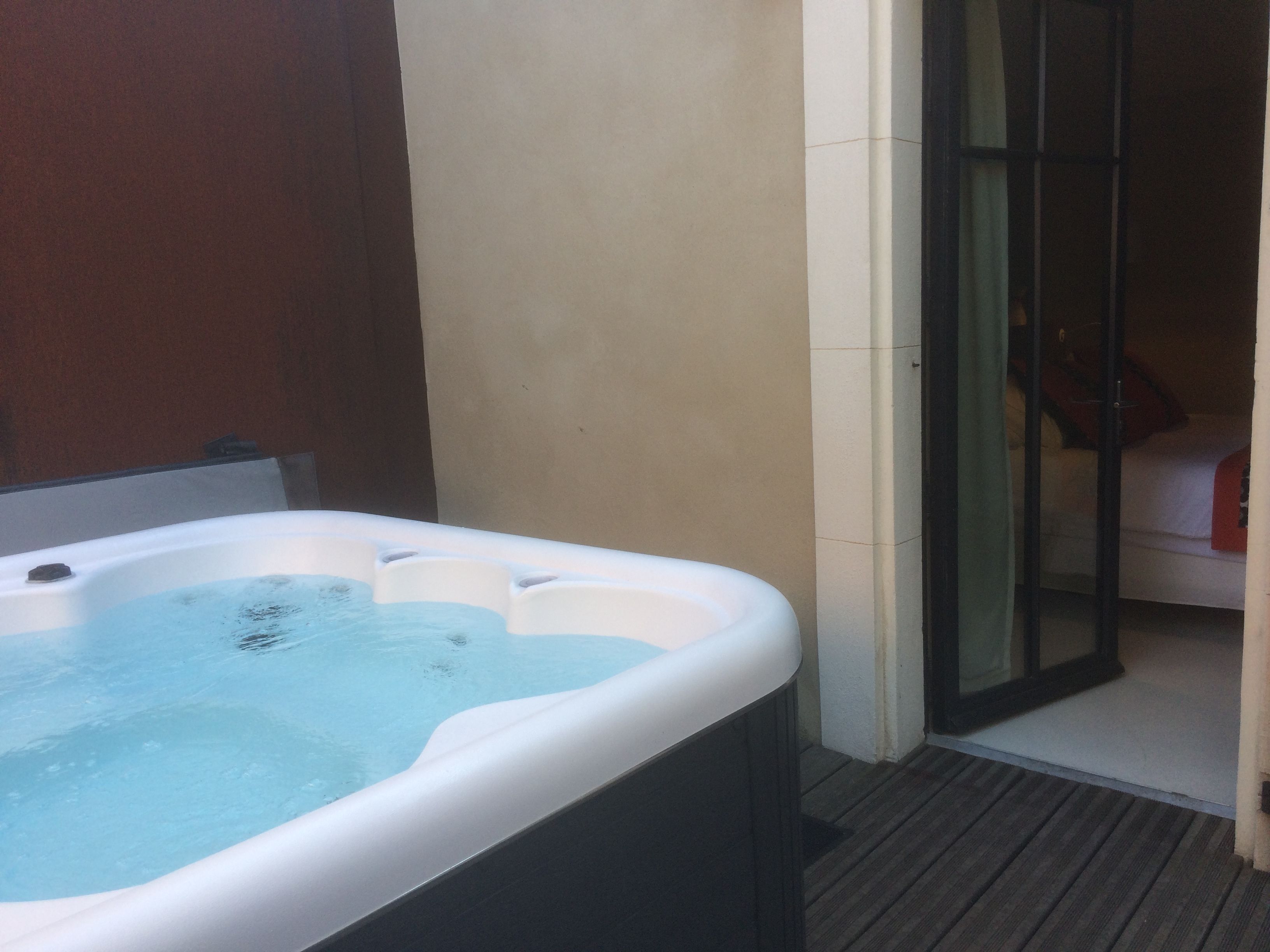 A hot tub with a wooden deck, a glass door leading to a bedroom with a bed and a red blanket.