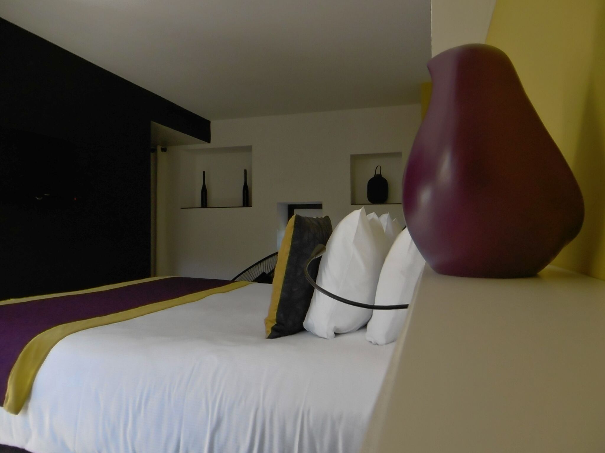 Une chambre d’hôtel avec un lit, un grand vase et une étagère remplie de bouteilles.