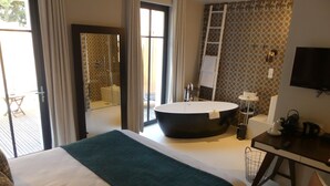 Une chambre d’hôtel avec un grand lit, une baignoire ronde, un bureau avec une chaise, une télévision et un balcon avec une table et des chaises.