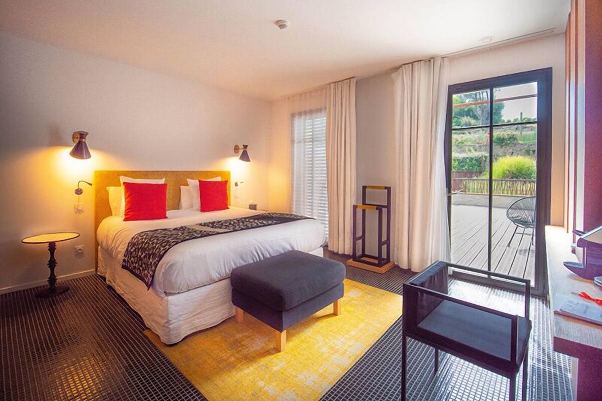 Habitación de hotel con cama, lámparas de noche, una mesita, un banco y vistas al exterior a través de amplios ventanales.