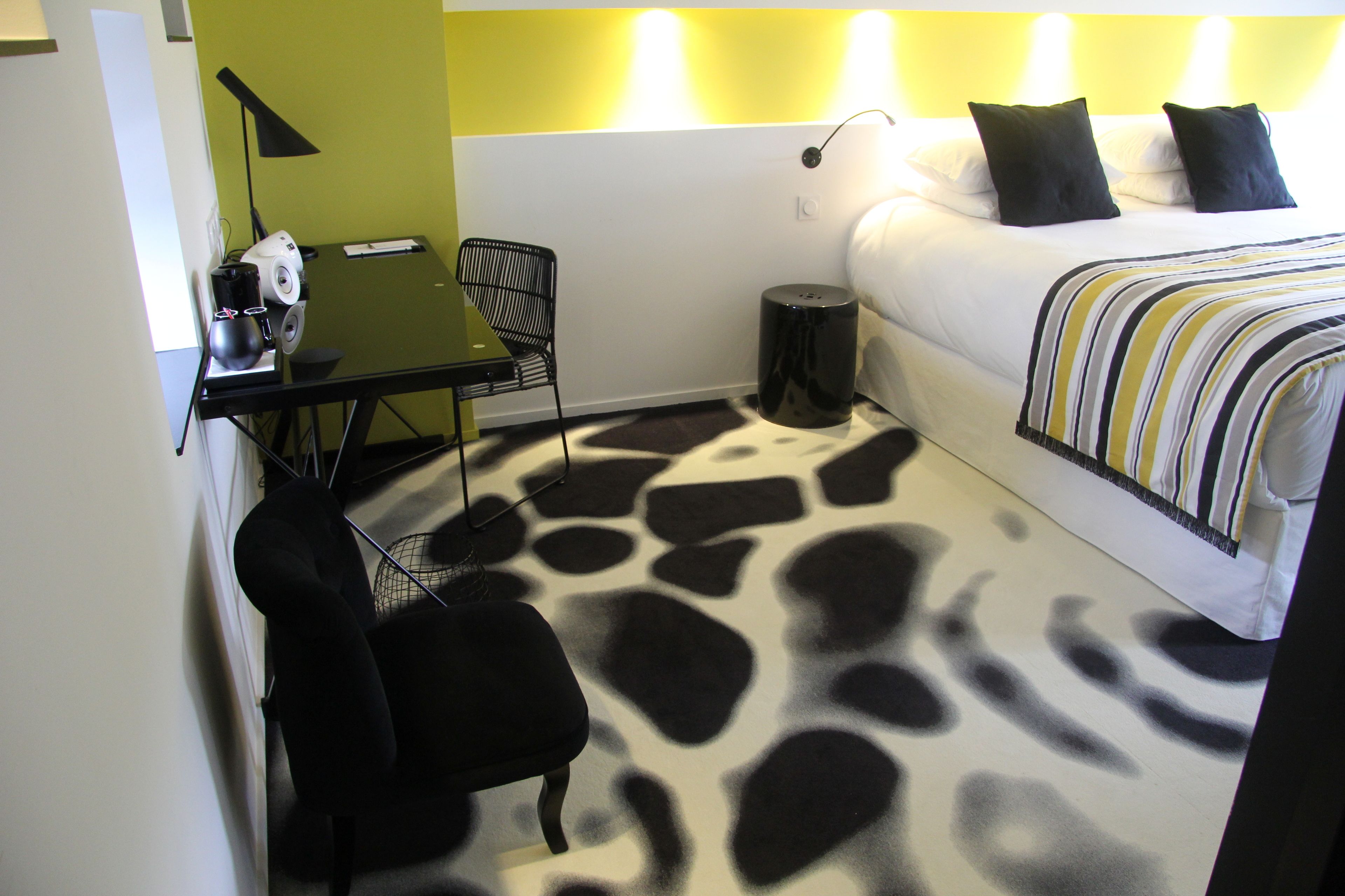 Quarto de hotel com cama, escrivaninha com cadeira, mesinha de apoio com abajur e tapete estampado com girafas.