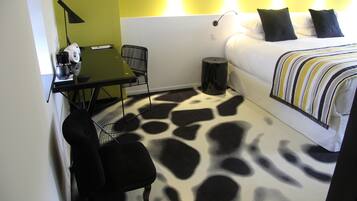 Quarto de hotel com cama, escrivaninha com cadeira, mesinha de apoio com abajur e tapete estampado com girafas.