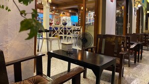 An indoor dining area with wooden tables and chairs, a fan, and a view into the restaurant through glass doors. - Steve Lady Hostel (Bangkok)