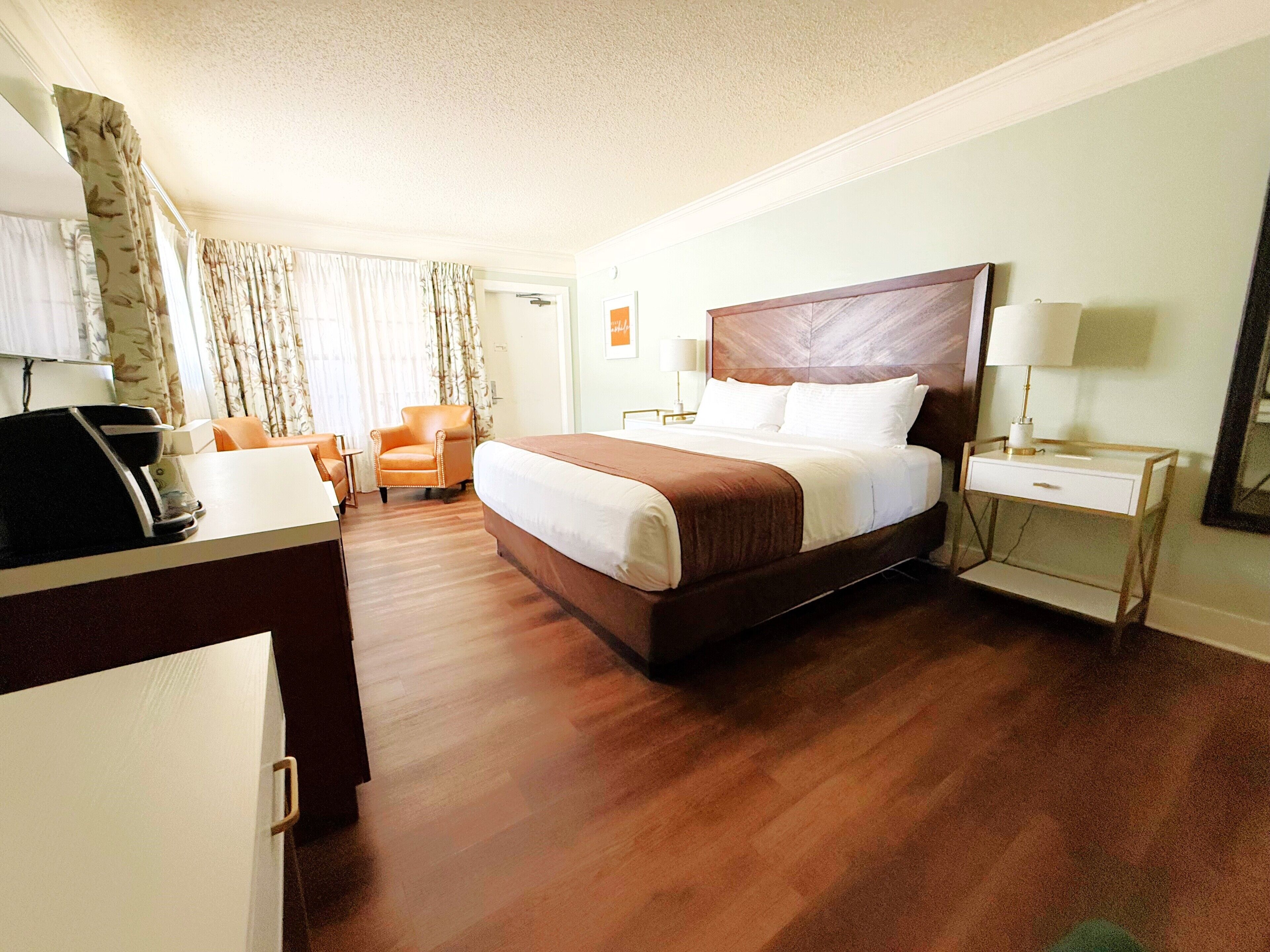 A hotel room with a large bed, a desk, a chair, a bedside table with a lamp, and a mirror.