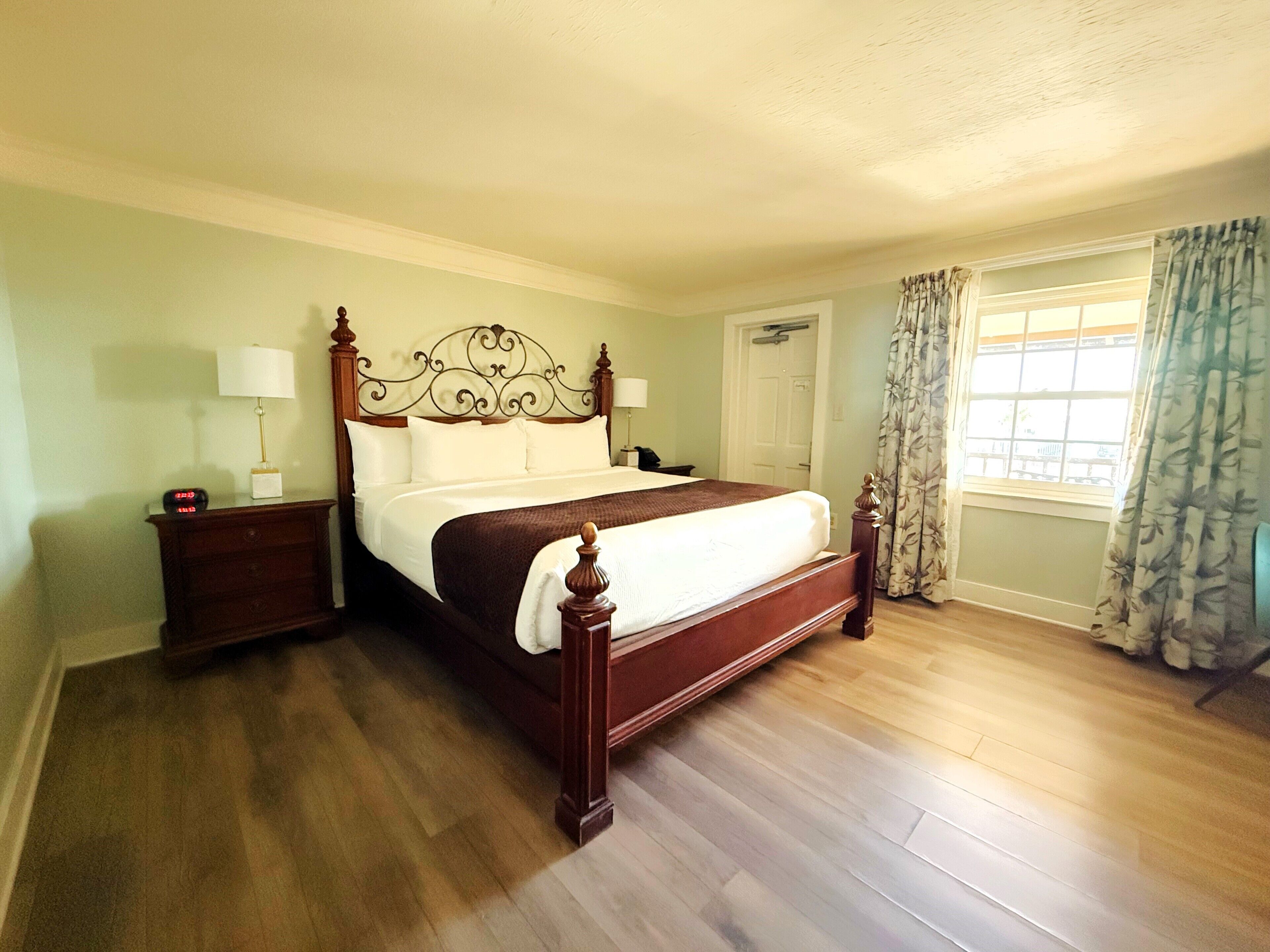 A neatly made bed with a wooden headboard, two bedside tables with lamps, a window with curtains, and a door in a room with wooden flooring.