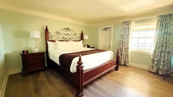 A neatly made bed with a wooden headboard, two bedside tables with lamps, a window with curtains, and a door in a room with wooden flooring.