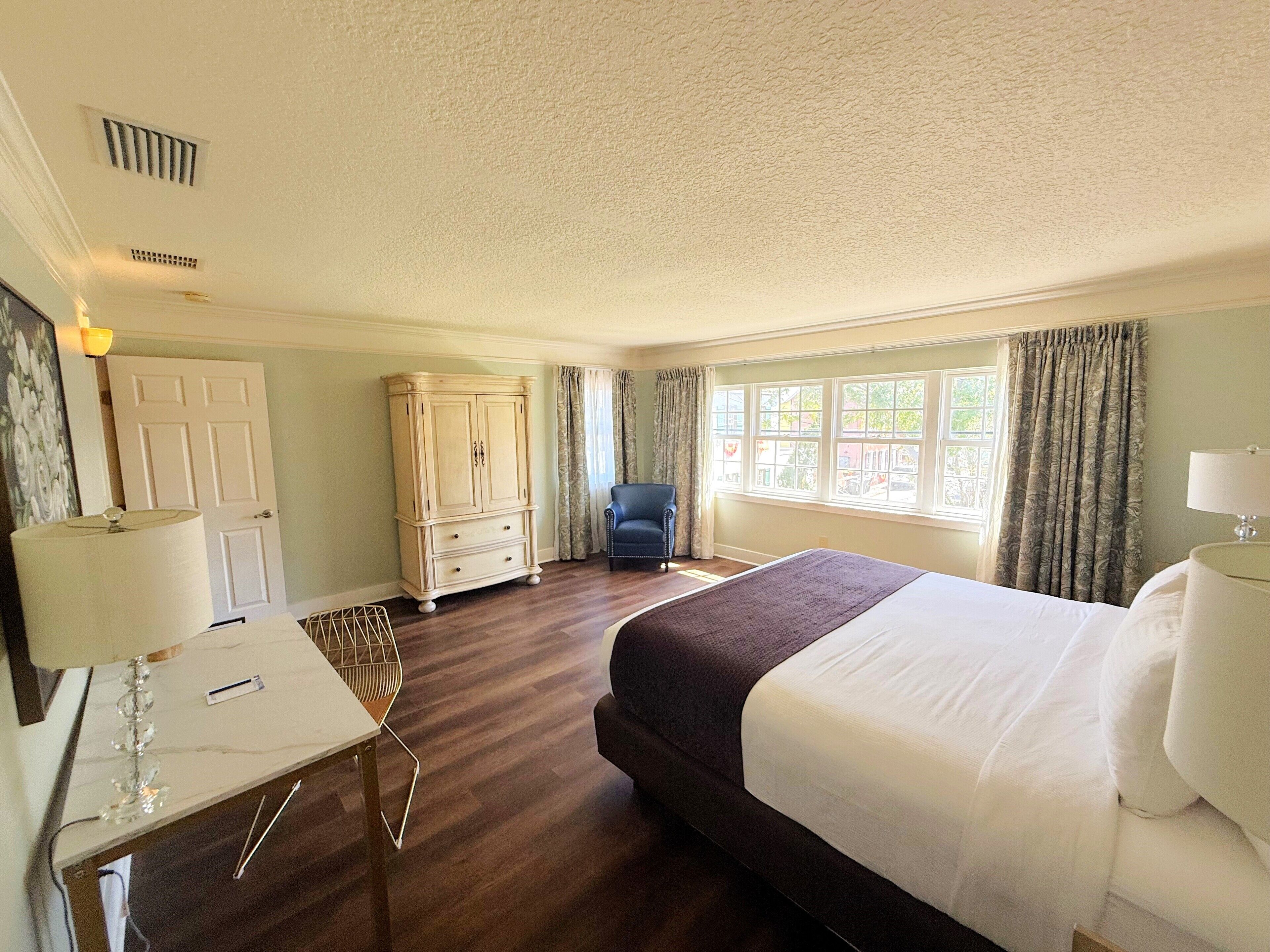 A hotel room with a large bed, a desk with a lamp, a chair, a wardrobe, and a window with curtains.