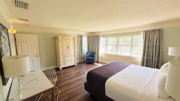 A hotel room with a large bed, a desk with a lamp, a chair, a wardrobe, and a window with curtains.
