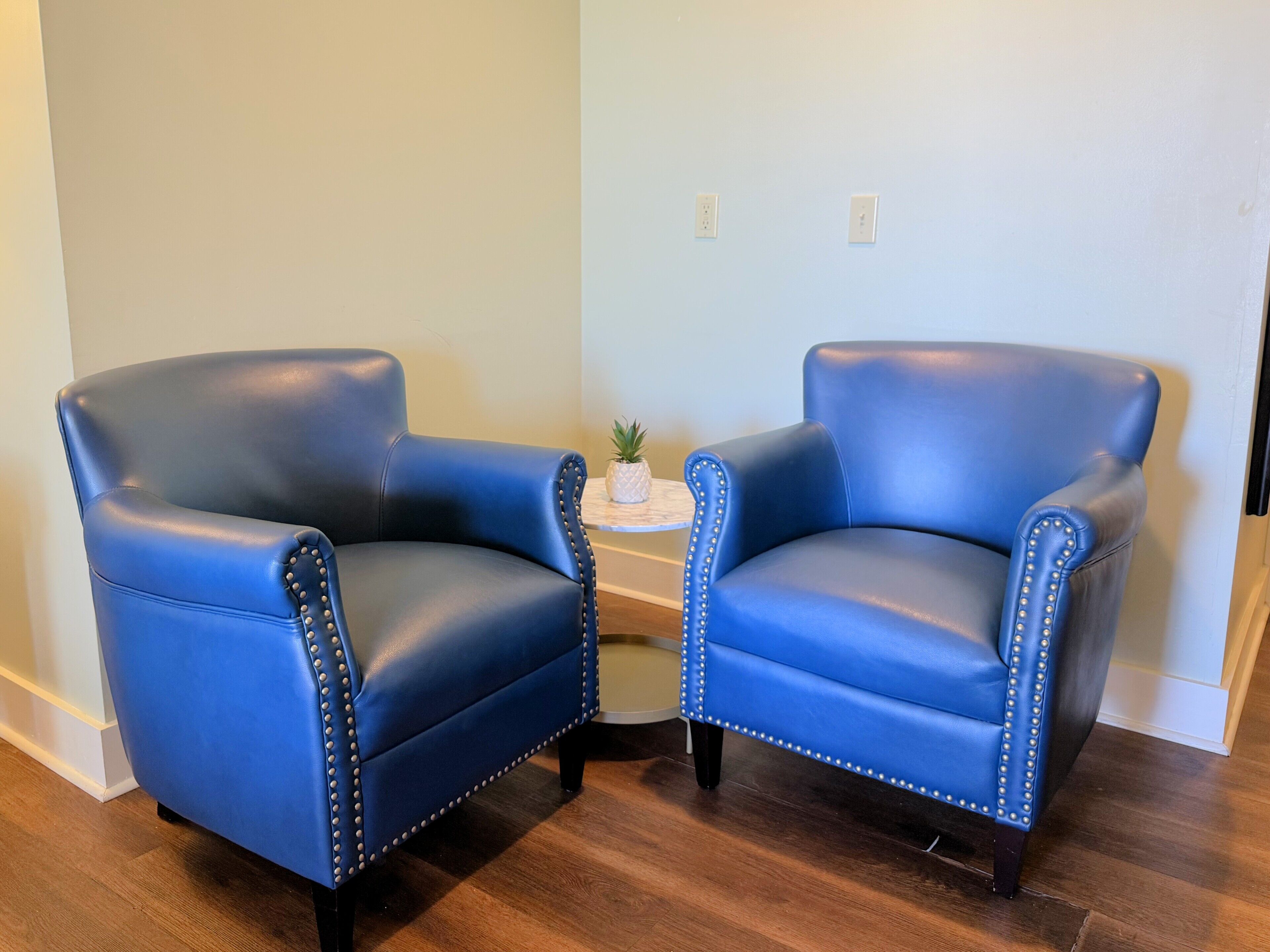 Two blue leather armchairs with metal studs, positioned on a wooden floor in a room with light-colored walls and a small round table with a plant.