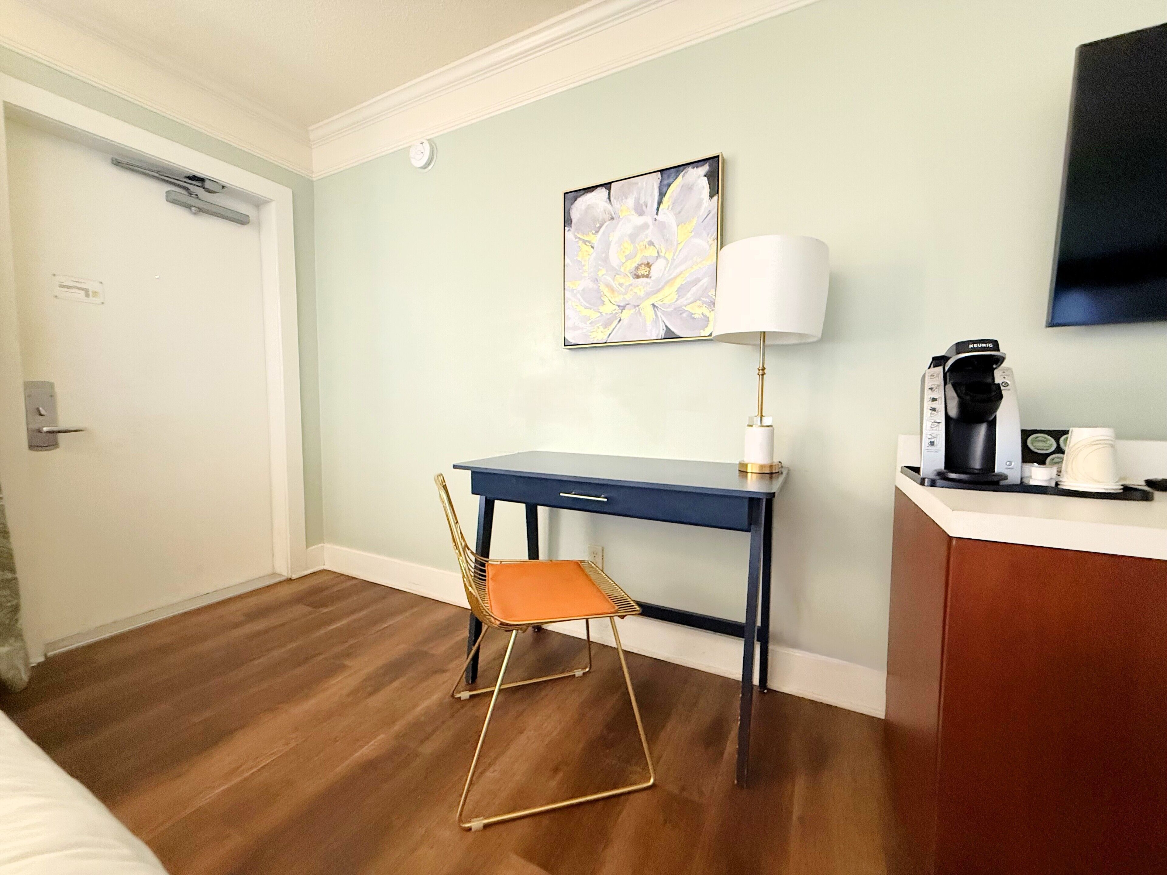 A hotel room with a desk, chair, coffee machine, and artwork on the wall.