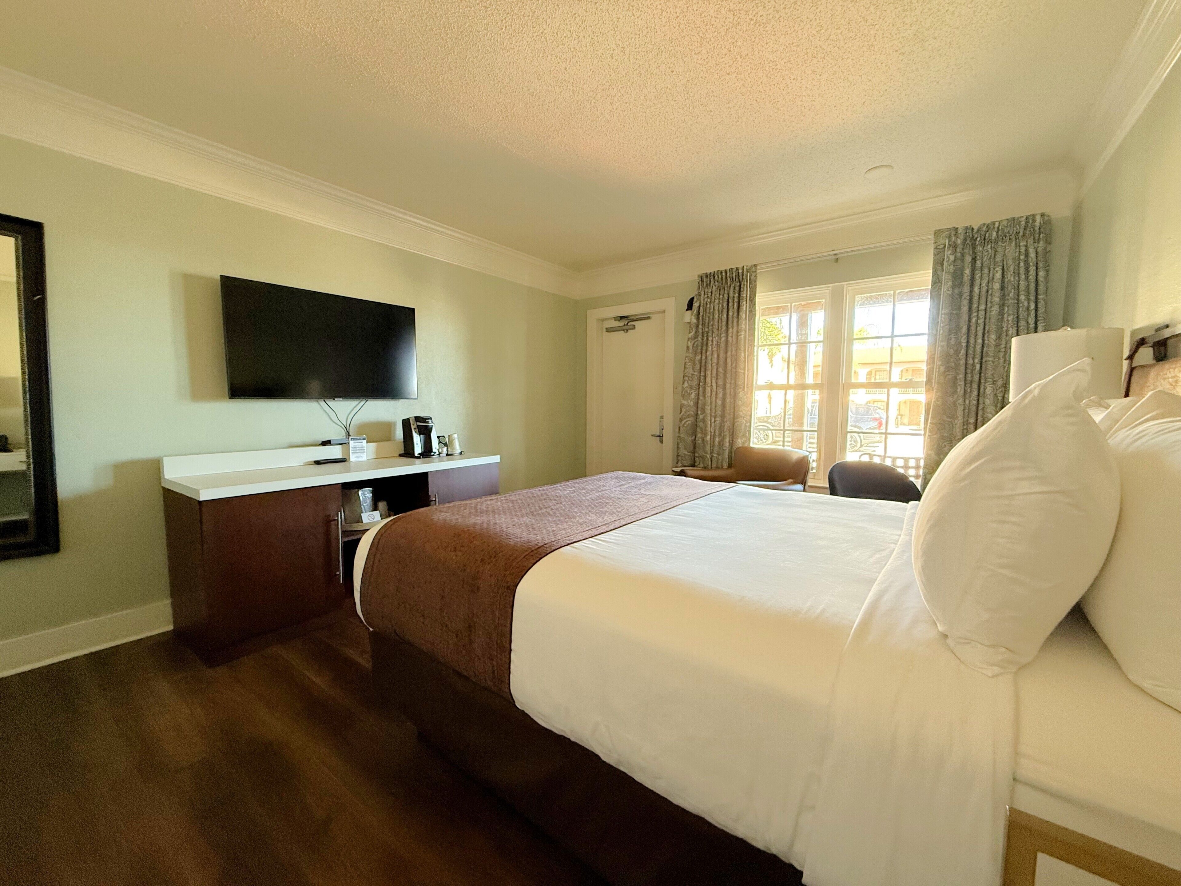 Habitación de hotel con cama, escritorio, televisor de pantalla plana, cafetera y ventana con cortinas.