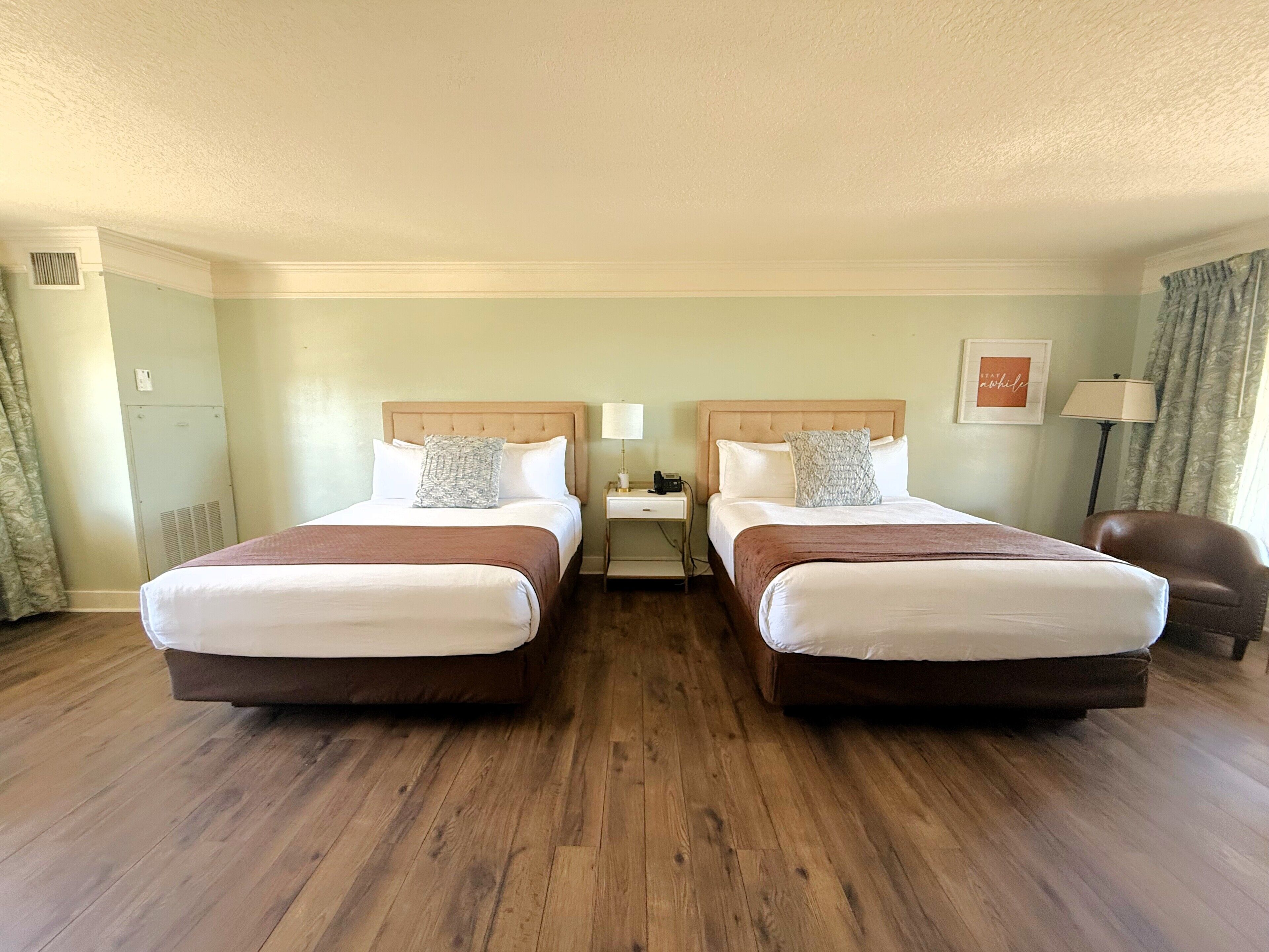 A hotel room with two beds, a nightstand, a lamp, a chair, and a framed picture on the wall.