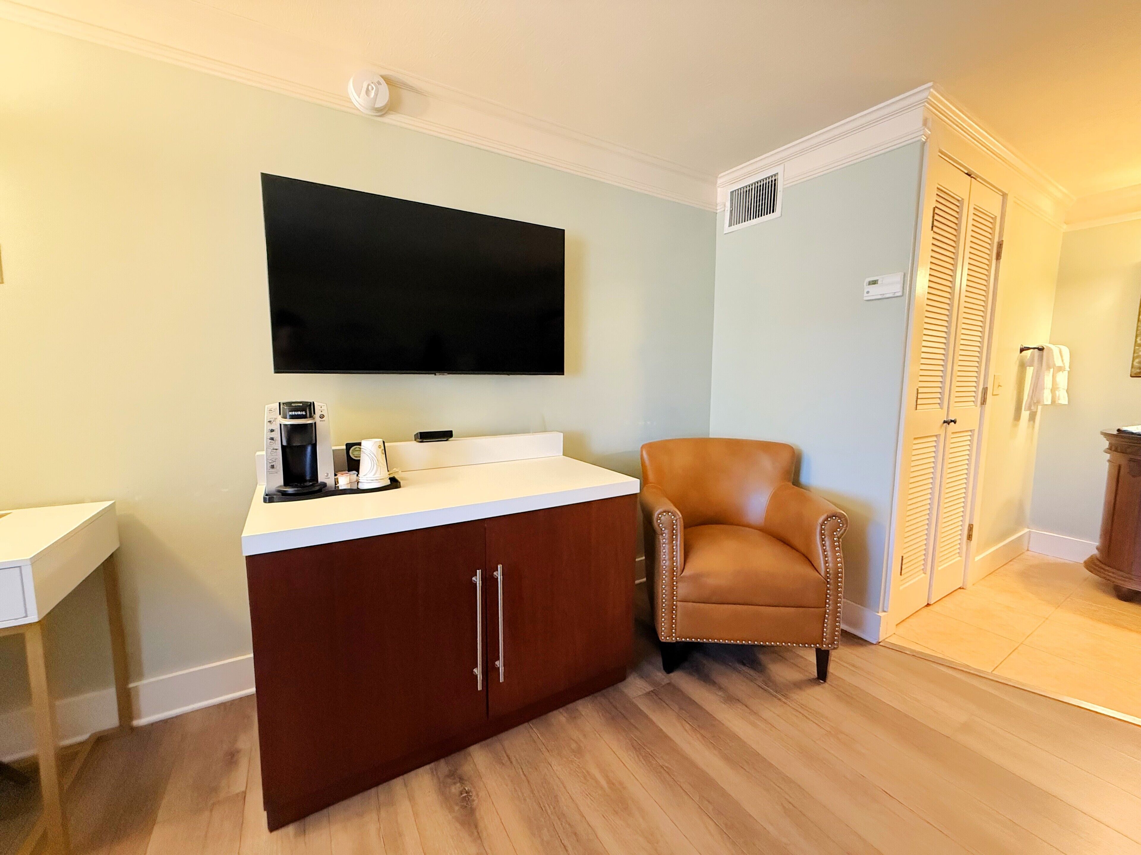 A hotel room with a wooden cabinet, a flat-screen TV mounted above it, a leather armchair, and a white bedside table.