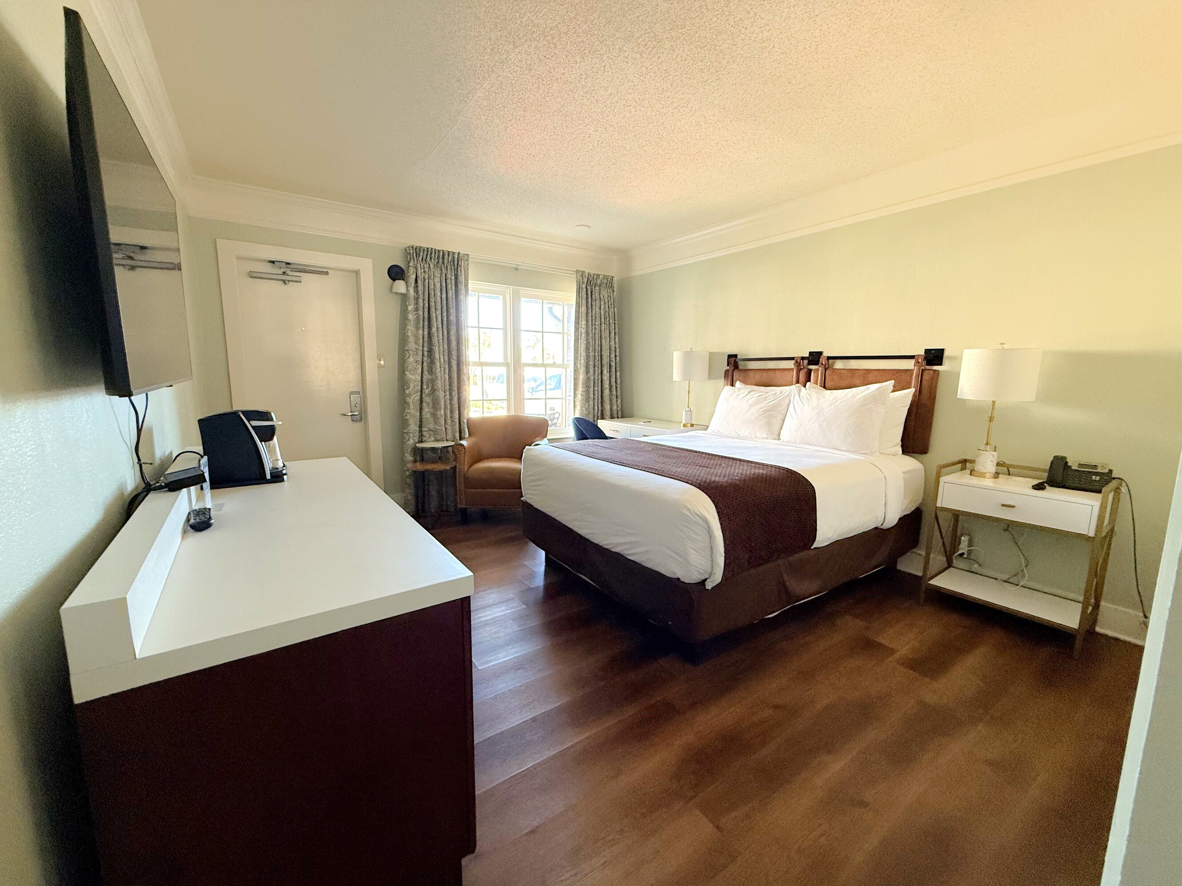 Ein Hotelzimmer mit einem großen Bett, einem Nachttisch mit Lampe, einem Sessel, einem Schreibtisch mit Computer und einem an der Wand befestigten Fernseher.