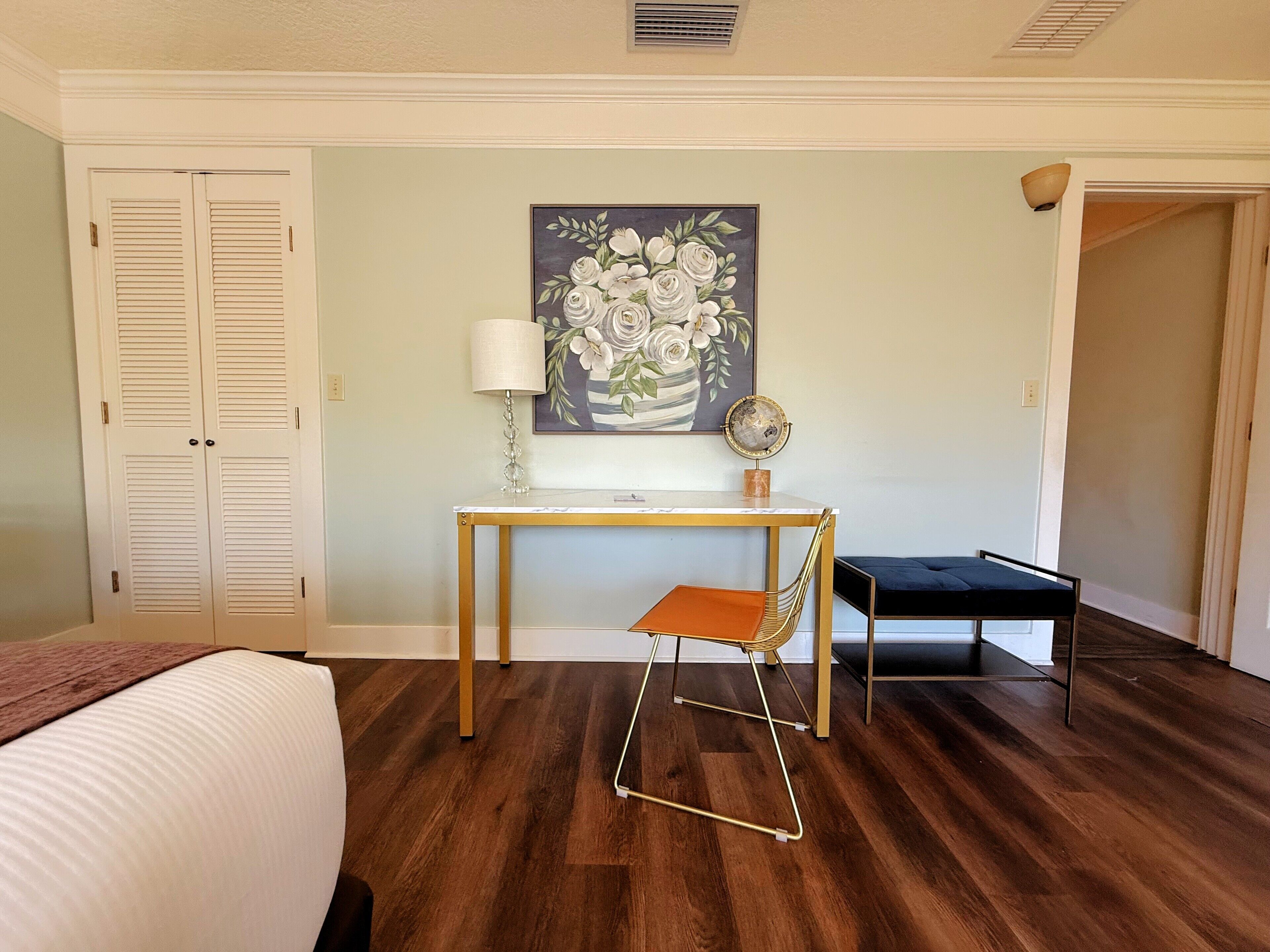 Una habitación de hotel con cama, escritorio con lámpara, silla, cuadro y espejo.