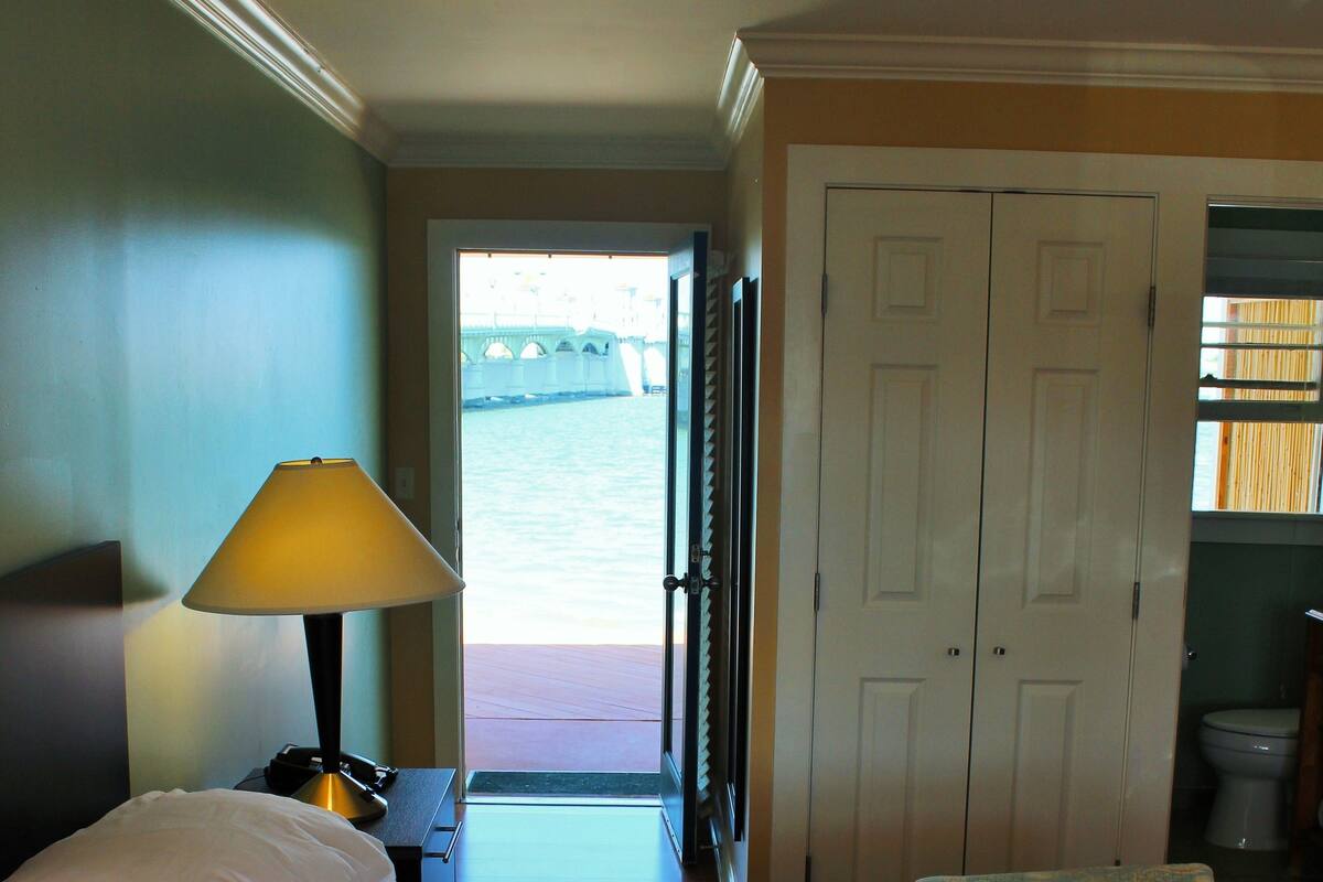 A hotel room with a bed, a nightstand with a lamp, a view of a bridge, and a door leading to another room.