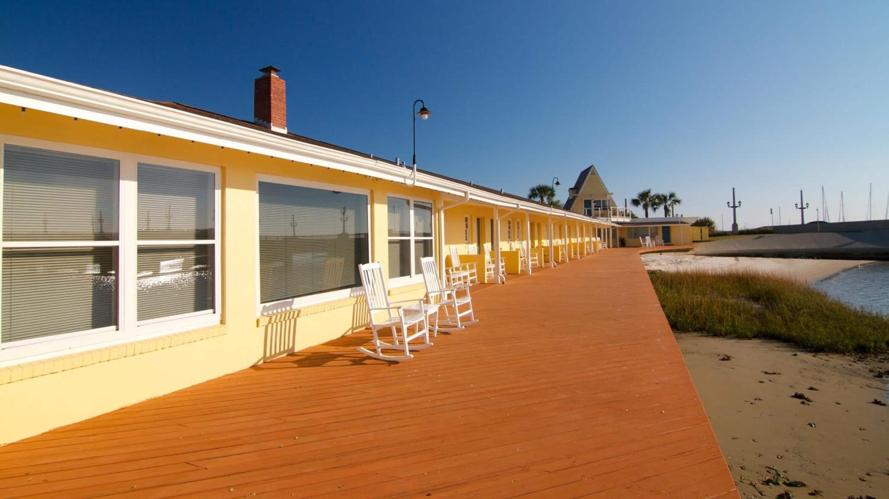 A beachfront hotel with a wooden deck, white rocking chairs, and a clear view of the ocean.