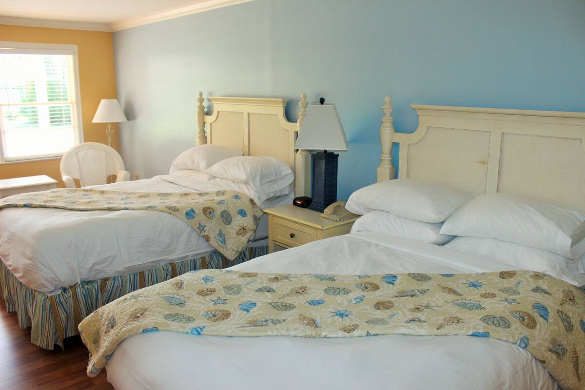 A room with two beds, each with a white bedspread and a patterned blanket. There is a nightstand with a lamp and a telephone between the beds.