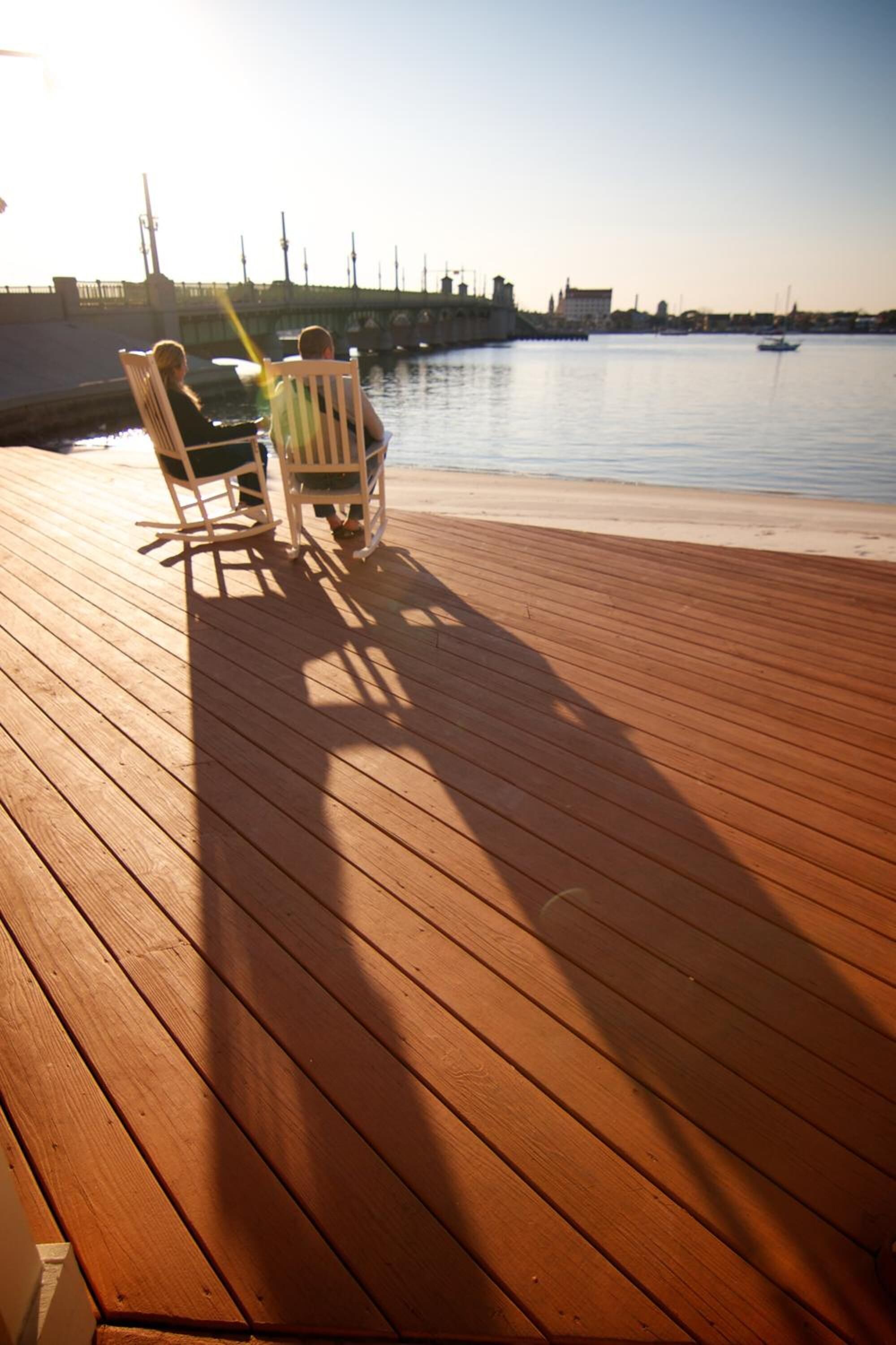 Una terraza de madera con dos mecedoras frente a un tranquilo paseo marítimo.