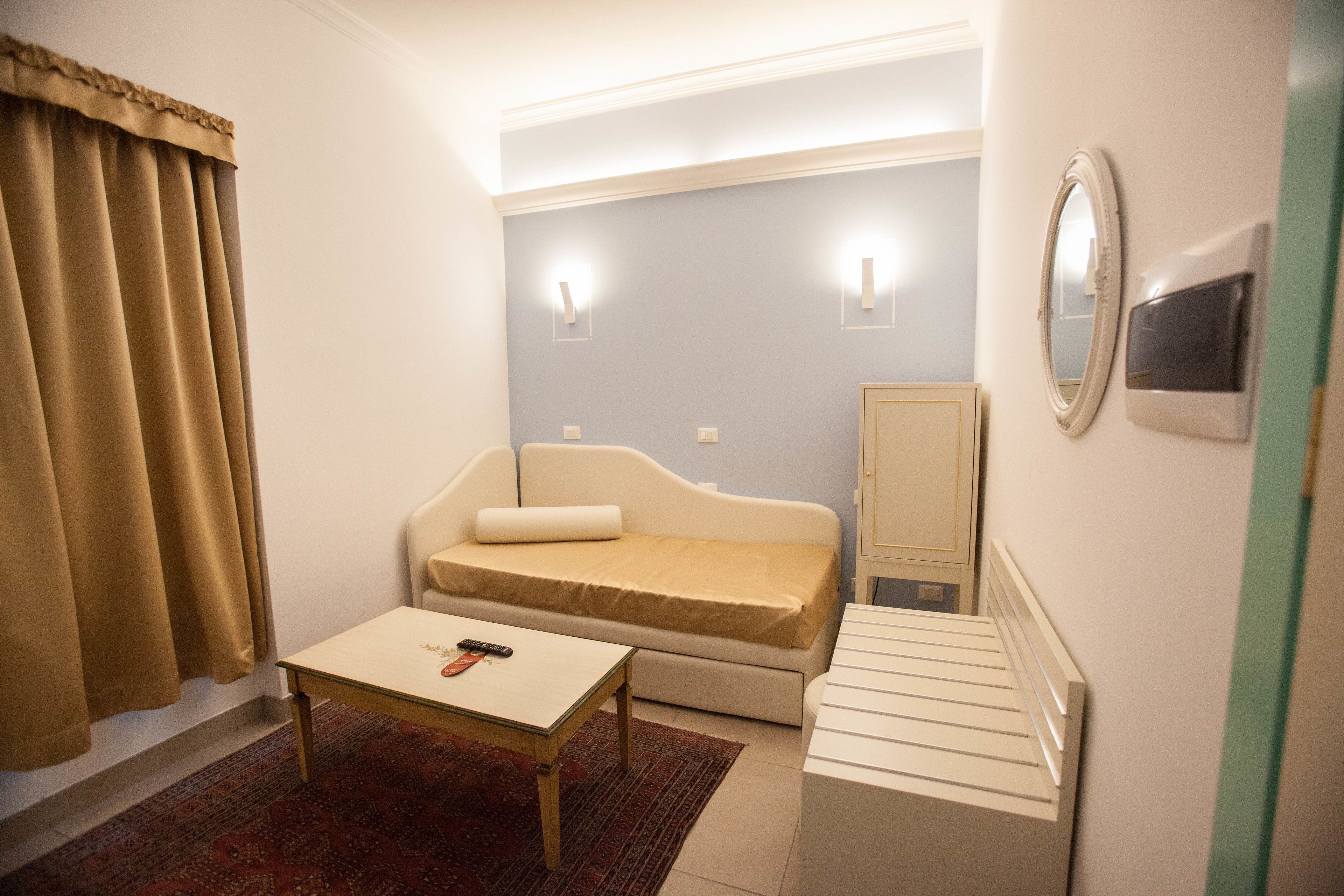 A small, well-lit room with a bed, a table, a mirror, and a radiator.