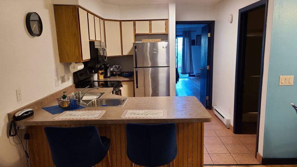 A kitchen with a stainless steel refrigerator, a microwave, and a sink. There are two blue chairs and a wooden counter.