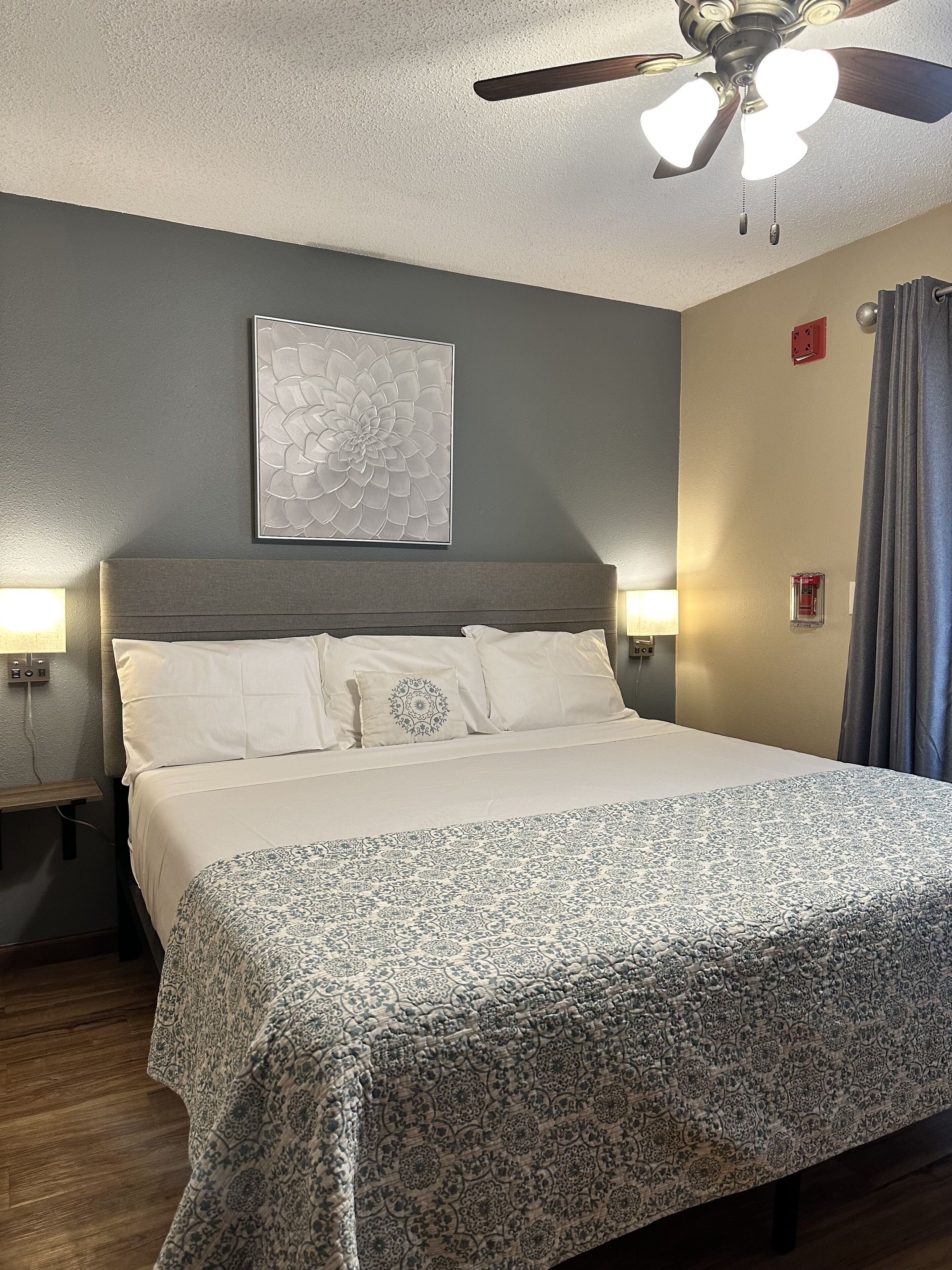 A neatly made bed with a patterned bedspread, a ceiling fan, a wall-mounted artwork, and a nightstand with a lamp.
