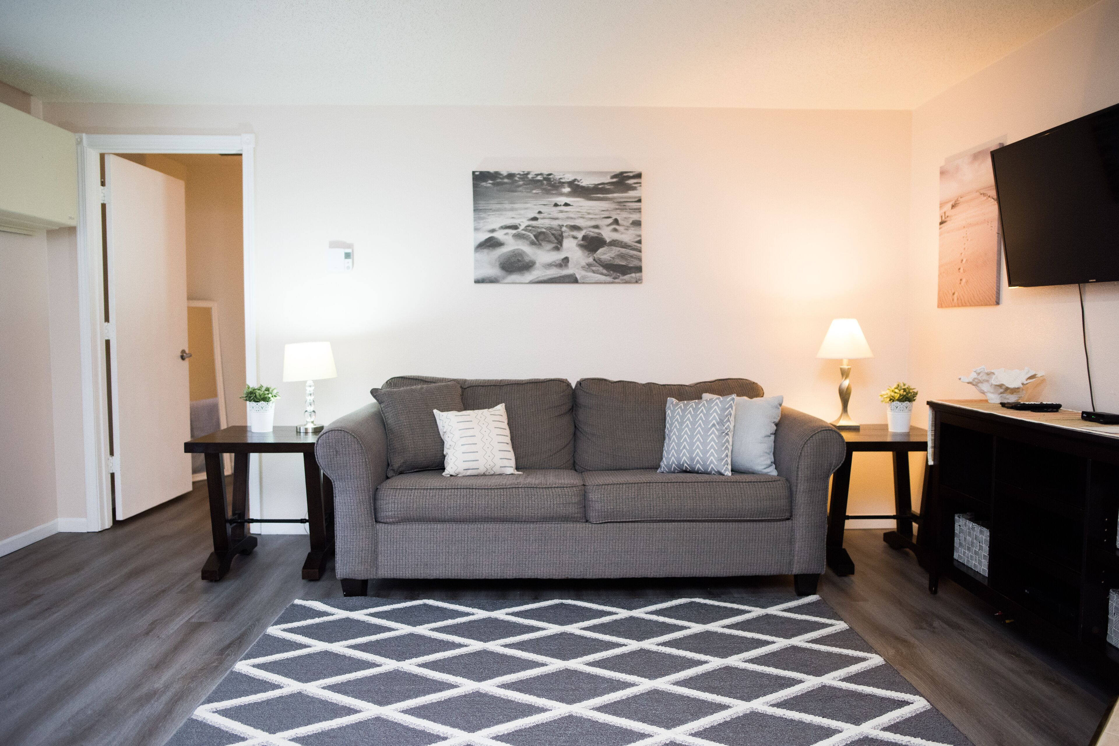 Un salon avec un canapé gris, deux tables d’appoint, une lampe, un téléviseur à écran plat et un tapis.