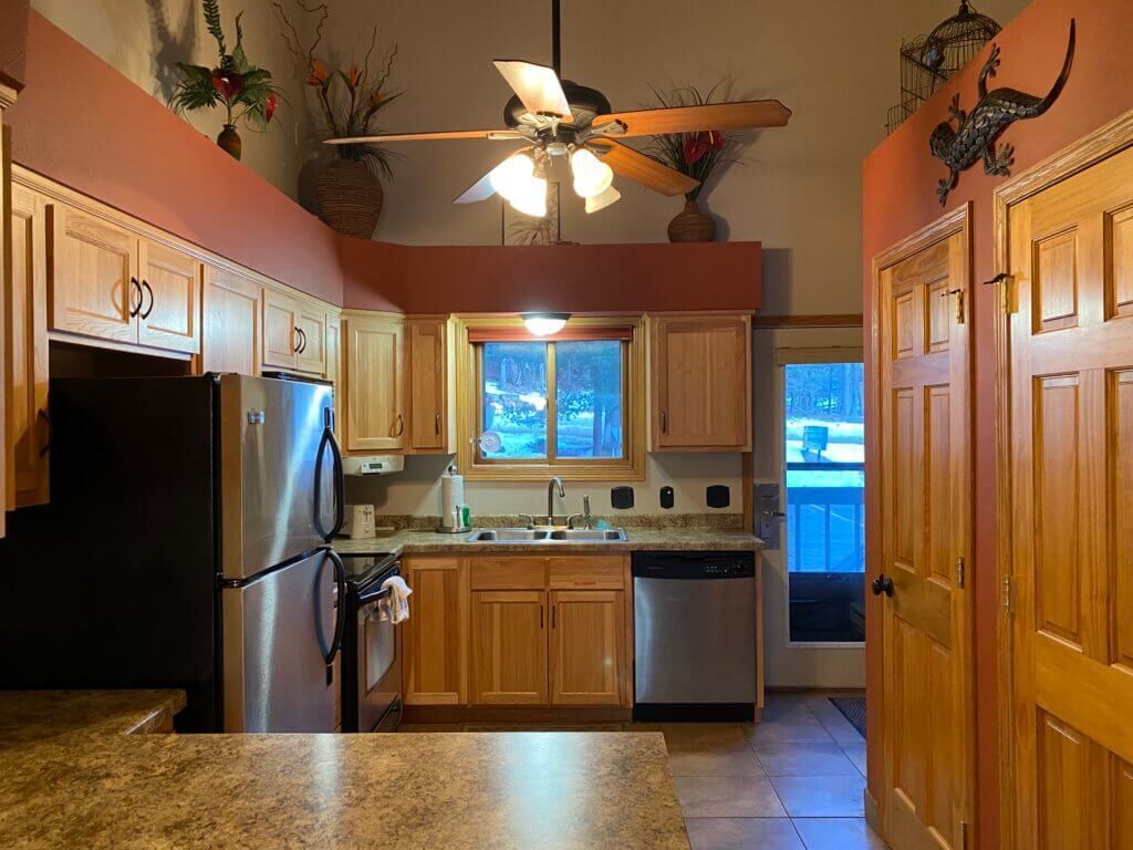 A kitchen with wooden cabinets, stainless steel appliances, a ceiling fan, and a window with a view of trees.