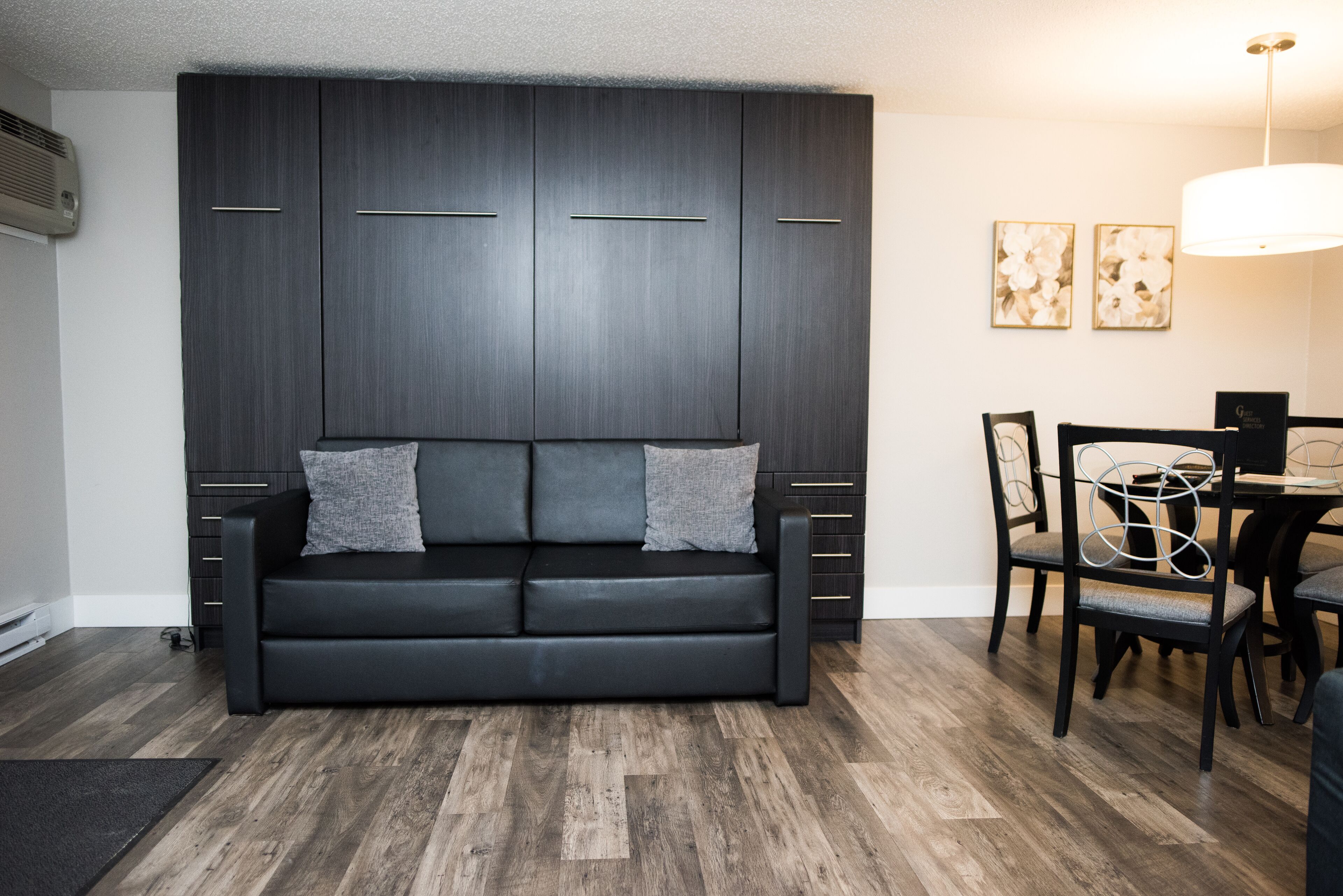 A modern living room with a black sofa, a dining table with chairs, and a dark wooden cabinet.